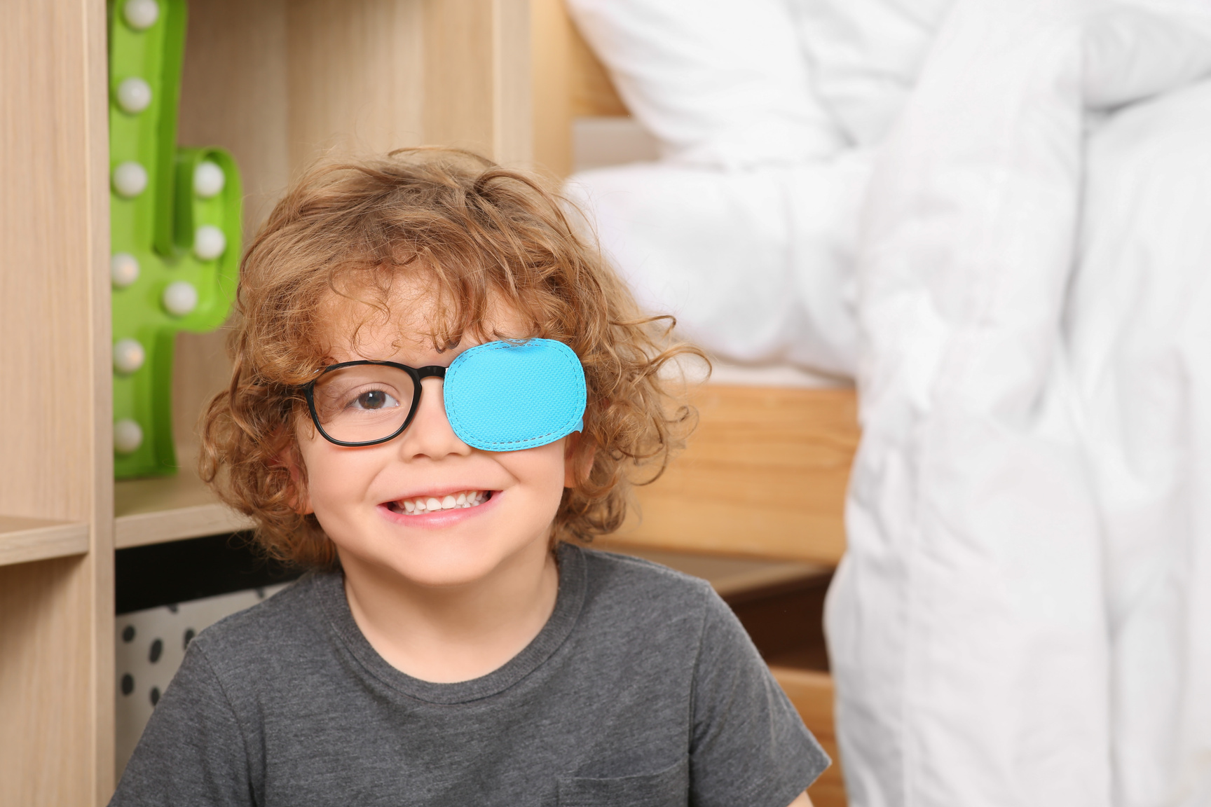 Happy Boy with Eye Patch on Glasses near Shelf Indoors. Strabism